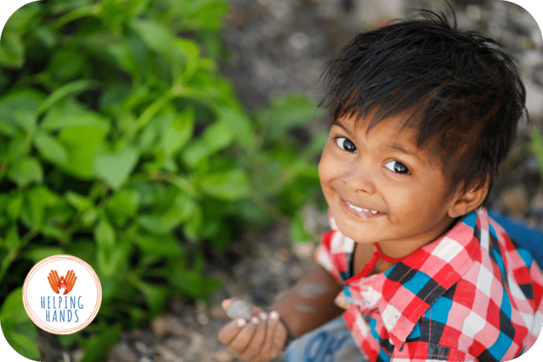 Petit garçon souriant en chemise rouge et blanche, assis près de plantes vertes, représentant l'impact positif d'un don pour les enfants démunis.