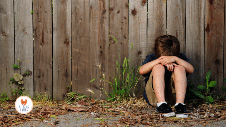 Enfant seul assis au sol contre une clôture en bois, tête baissée sur les bras – symbole de solitude et d’enfants oubliés, avec le logo de Helping Hands