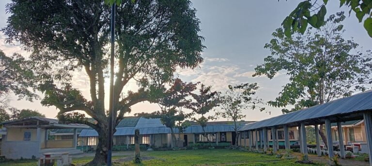 Vue du bâtiment principal de l’école publique de Limbauan, aux Philippines, au lever du soleil, entouré d’arbres et de structures de plein air.