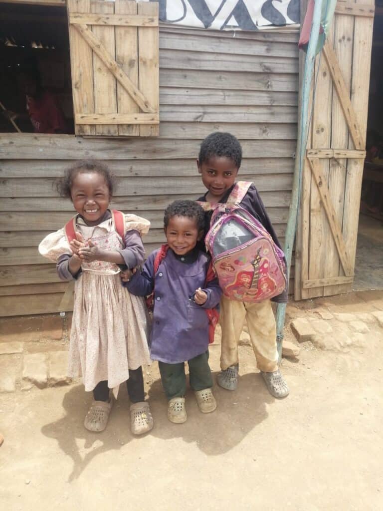 Trois enfants malgaches souriants devant une salle de classe en bois à Antsirabé, portant leurs sacs à dos. Photo illustrant l'action de Helping Hands pour soutenir l'école et offrir de meilleures conditions de scolarisation.