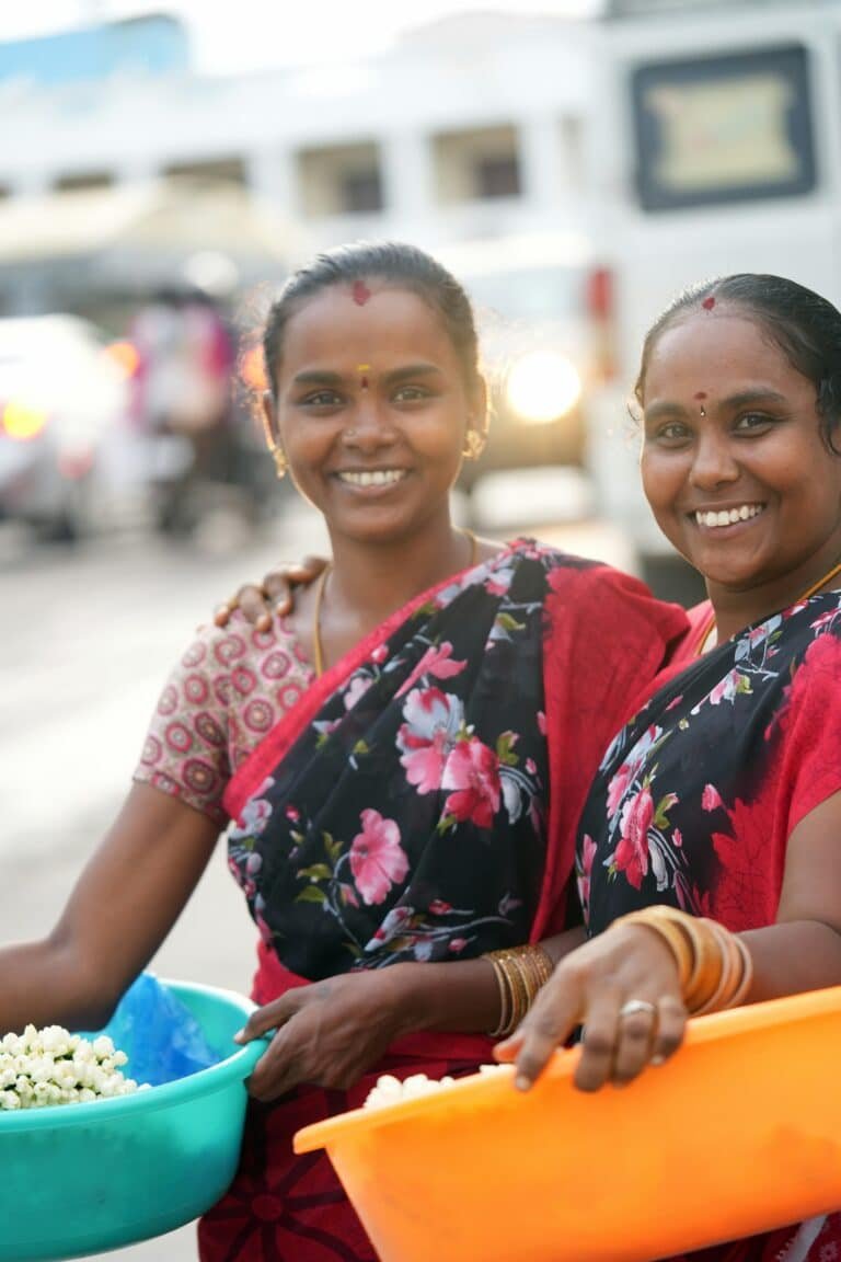 Deux femmes souriantes en sarris floraux, portant des bassines de fleurs